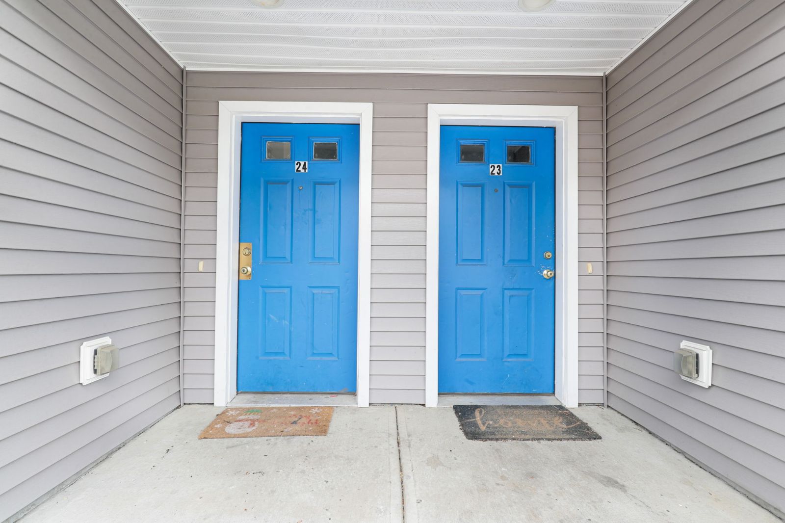Two blue front doors with doormats under a covered entry