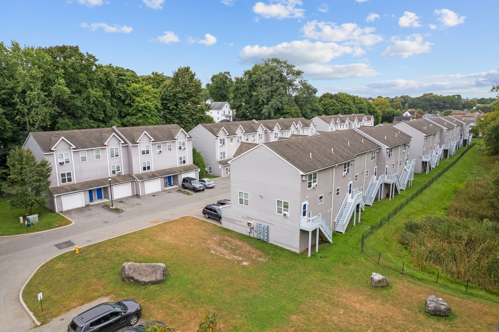 Low aerial view of the townhomes showing rear stairs and decks beside a lawn and a fenced green edge